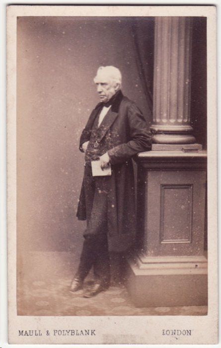 Full-length studio photo of an elderly William Fairbairn in a dark coat, standing beside a column