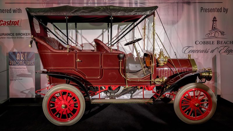 Red early touring car with brass lamps and spoked wheels on display at an auto show